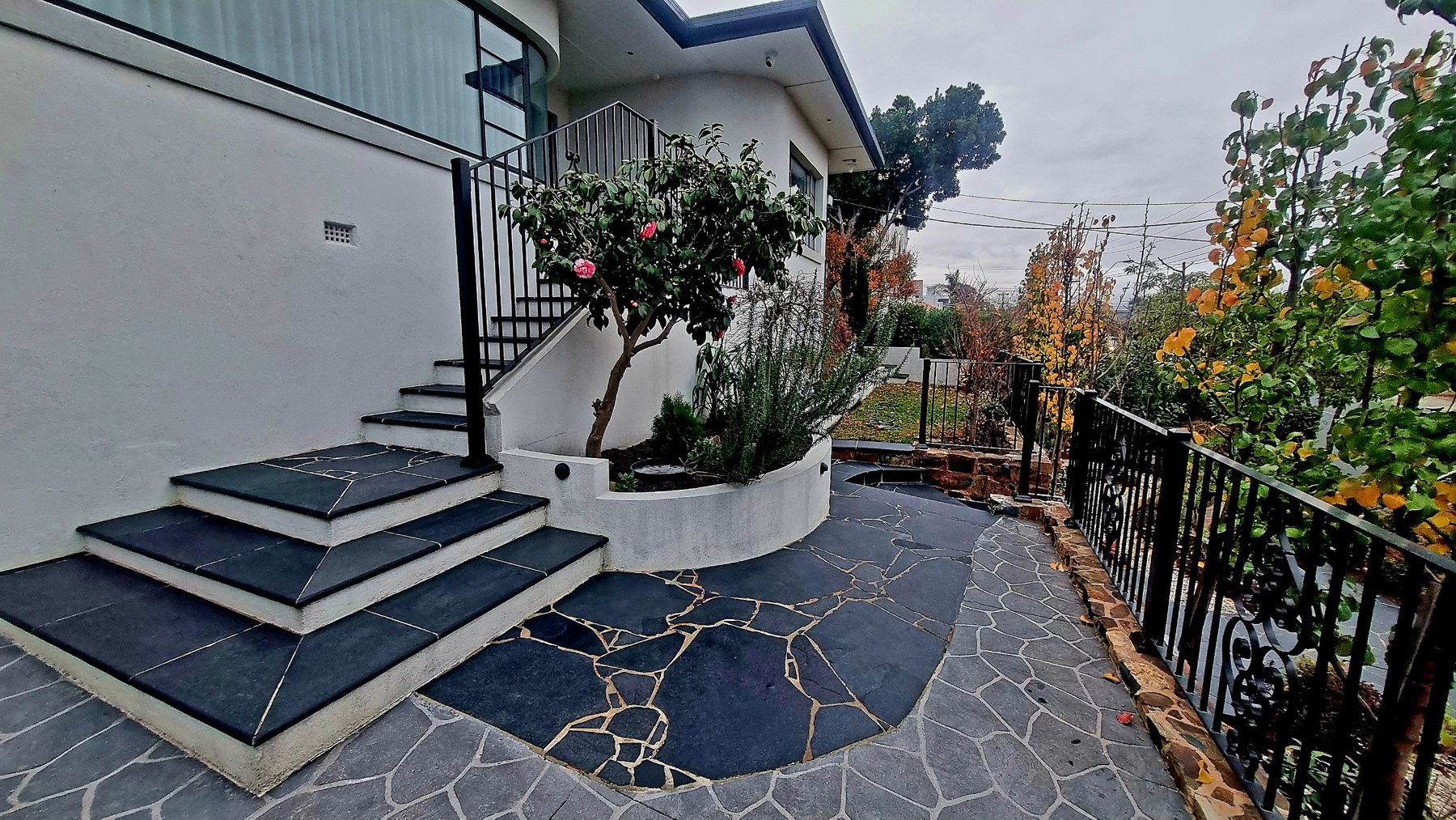 Black stone steps, curved planter and crazy paving along the side of the house
