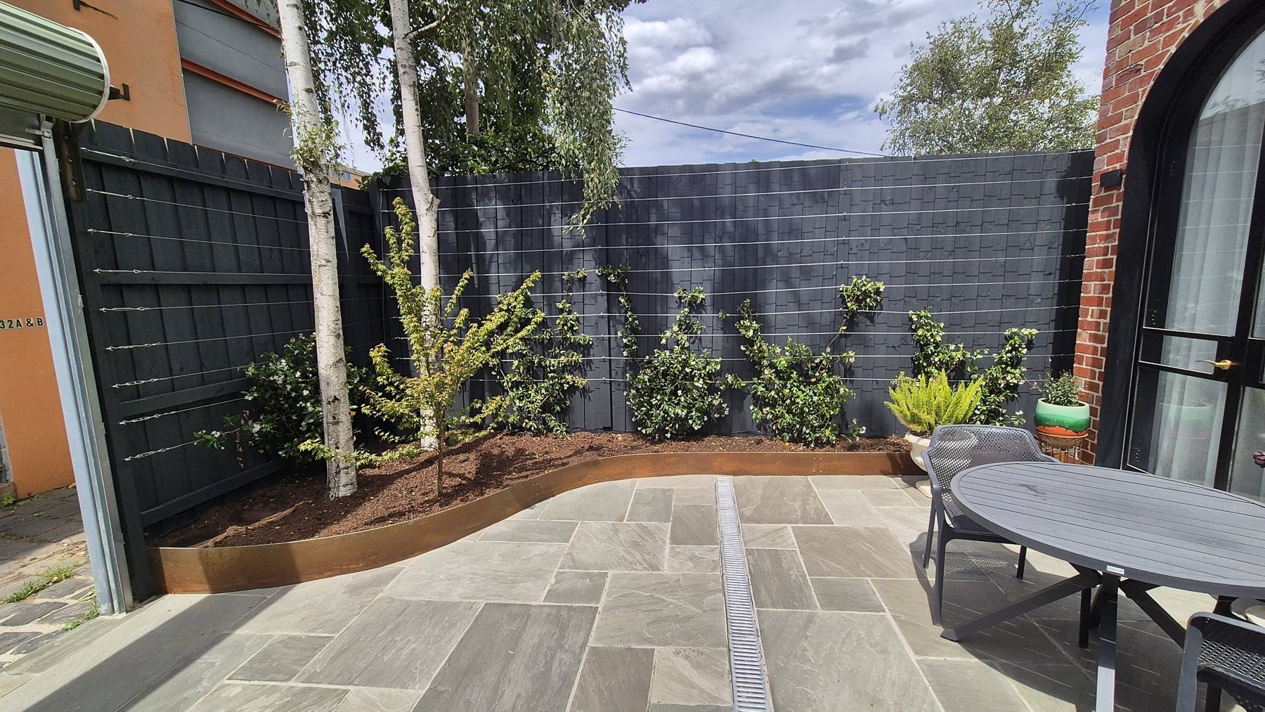 Curved Corten garden bed with silver birches and star jasmine on wires along a charcoal fence
