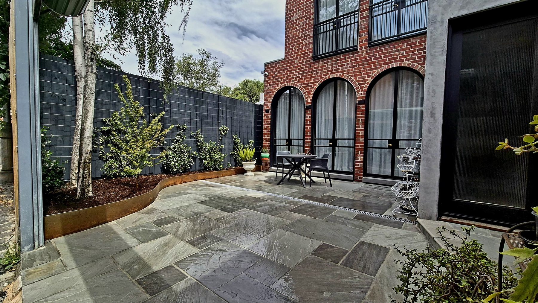 Grey stone paving courtyard with a curved Corten bed, silver birches and arched brick windows