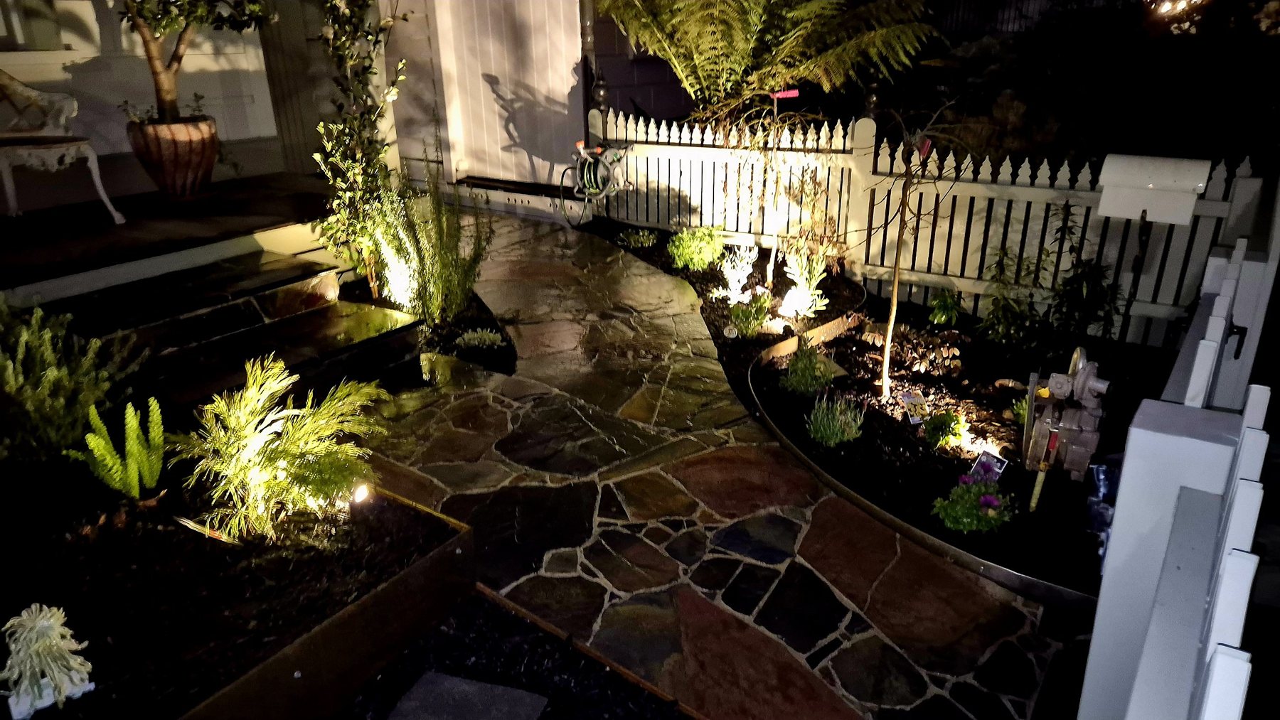 Slate crazy paving and Corten edged garden beds lit at night beside the picket fence