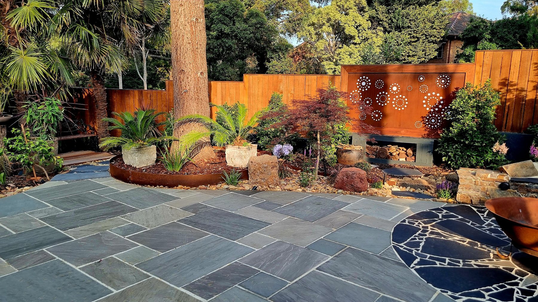 Corten screen with circle cut-outs above a log store, with Corten edged garden and stone paving
