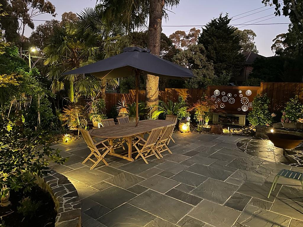 Dining terrace at night with uplit palms, lit garden and the Corten screen glowing