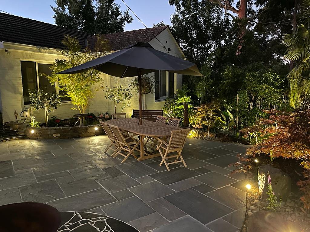 Courtyard dining terrace under an umbrella, with lit stone walls and garden at night