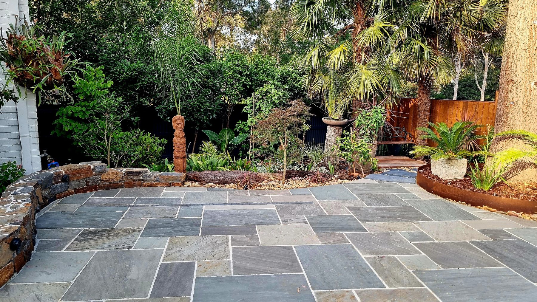 Grey stone paving curving past a carved timber totem, low stone wall and lush planting
