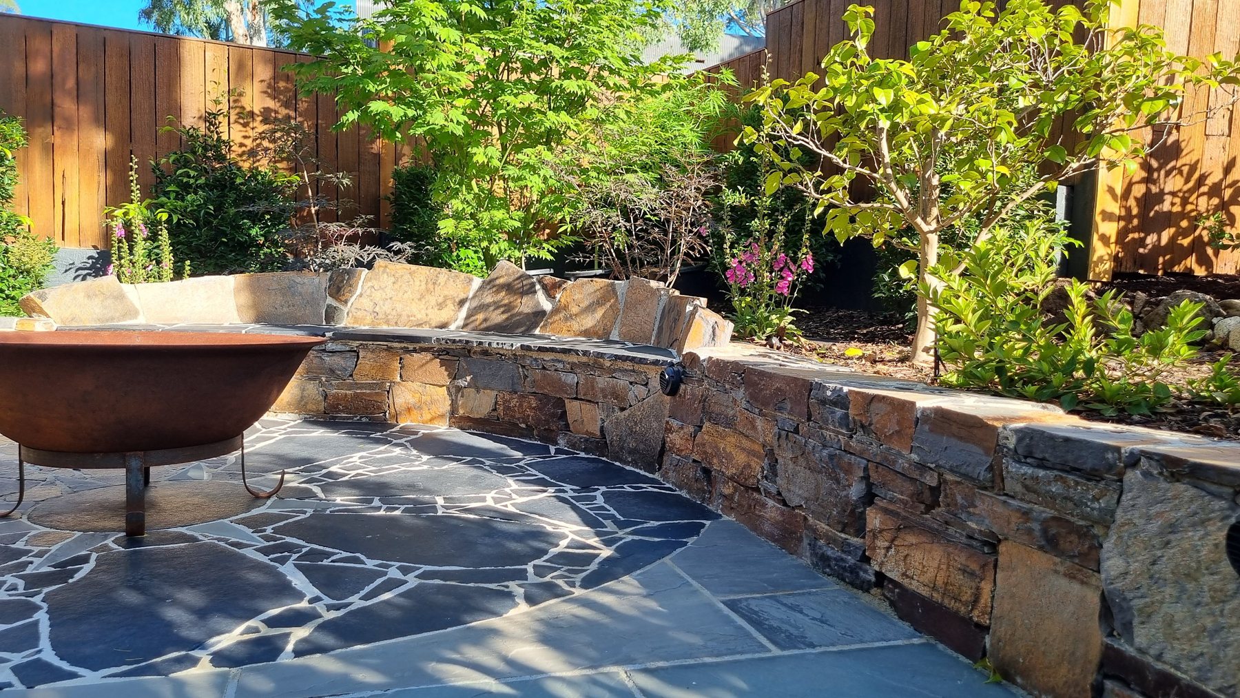 Curved stacked stone seat wall and fire pit on black crazy paving in sunlight