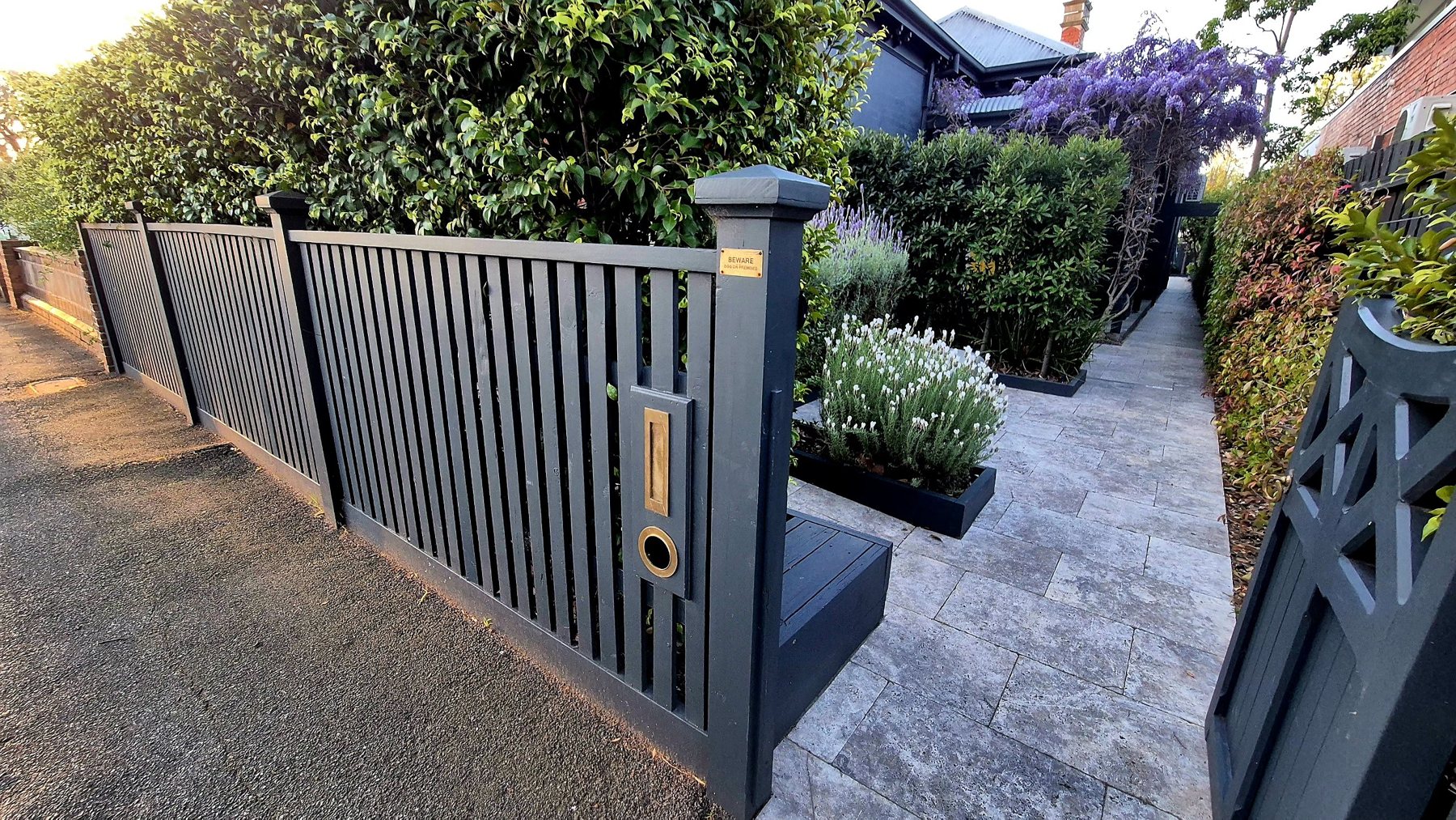 Charcoal slat front fence with brass letterbox, travertine path and wisteria beyond