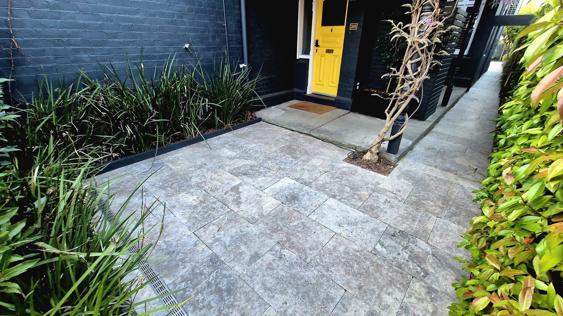 Travertine paving to a yellow front door, with a strip drain and clipped planting