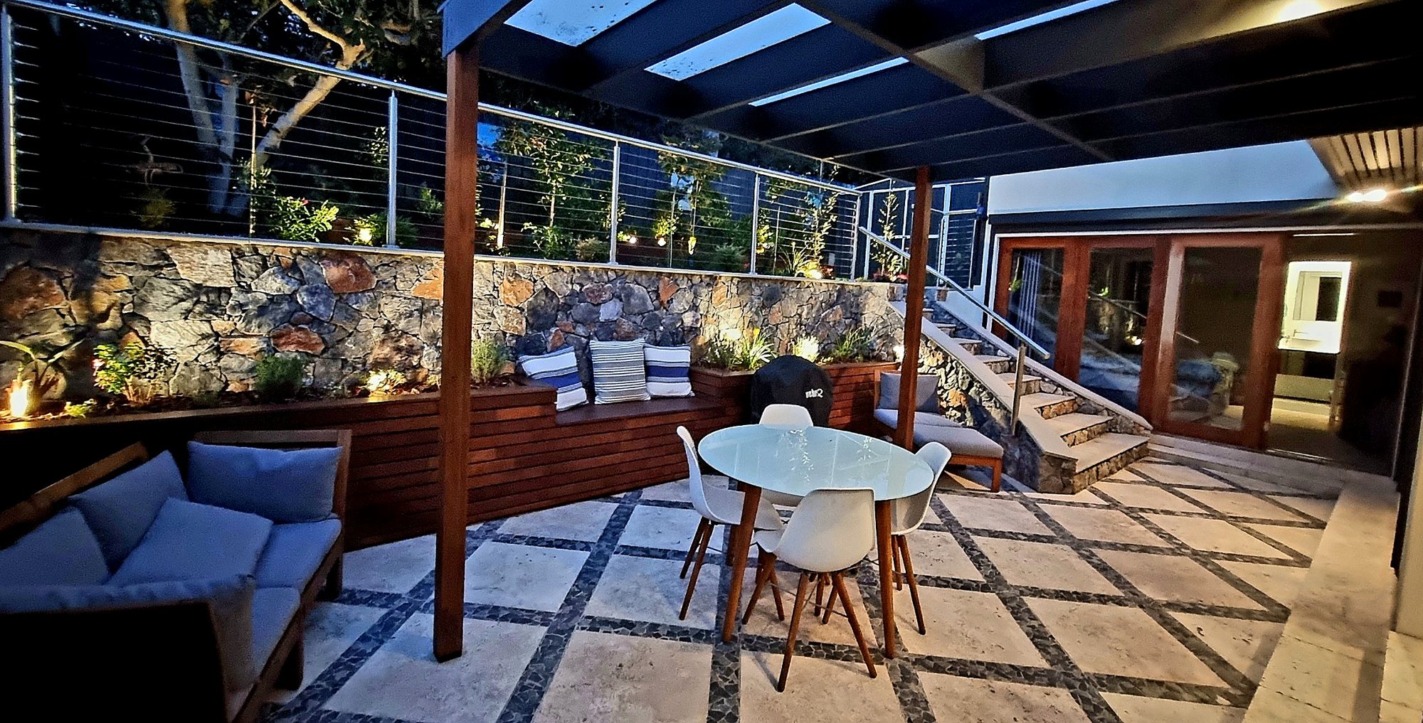 Courtyard at dusk with a natural stone retaining wall, timber bench planters, a timber pergola and lit garden beds above