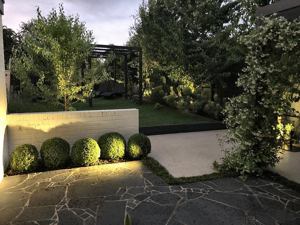 Lit buxus balls and crazy paving at dusk, with the pergola beyond